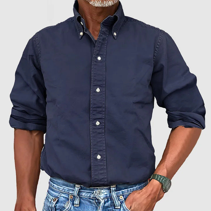 A man wearing a navy button-down shirt with the sleeves rolled up to the elbows, paired with blue jeans.