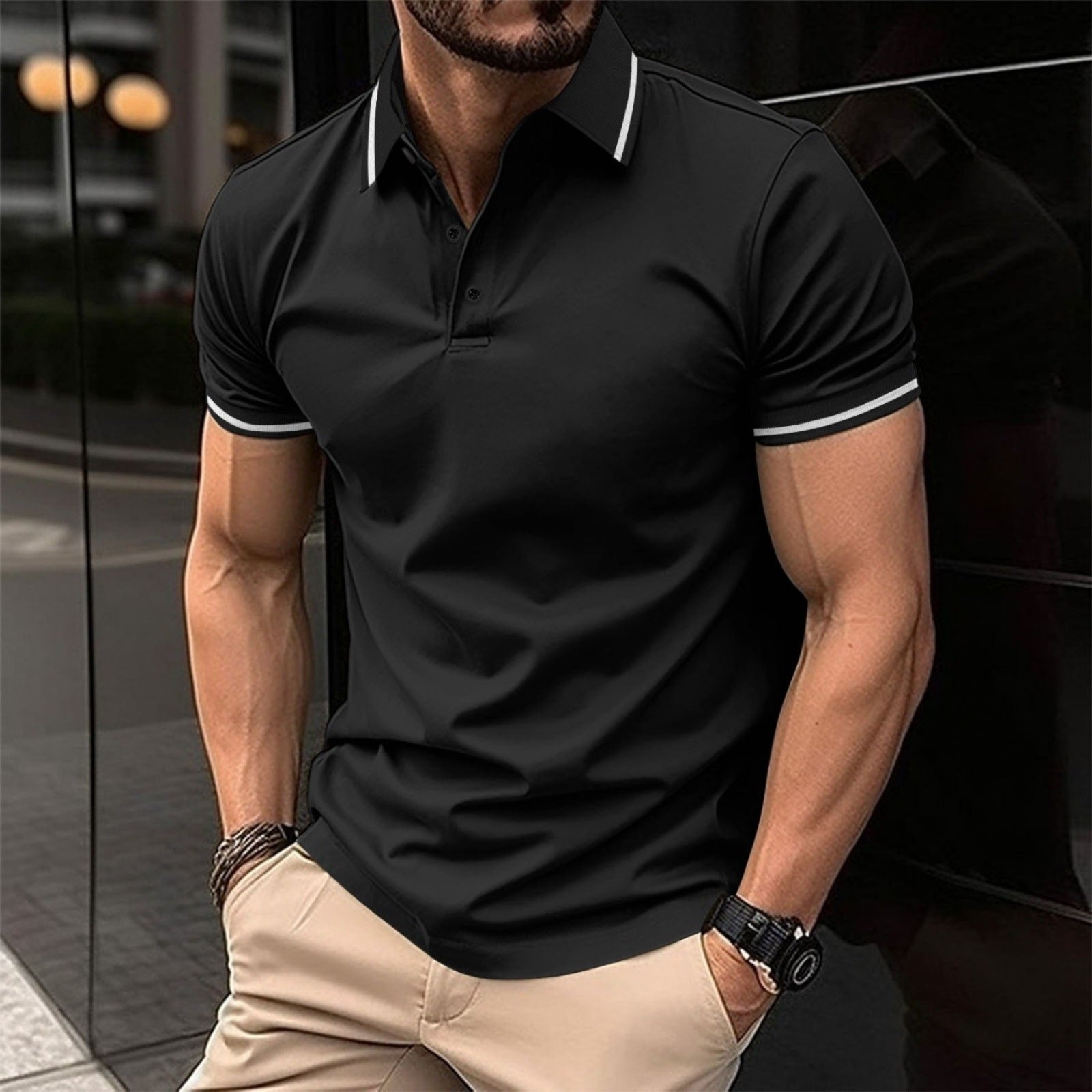 A man wearing a black polo shirt with contrast trim around the collar and sleeves, standing in a reflective indoor setting.